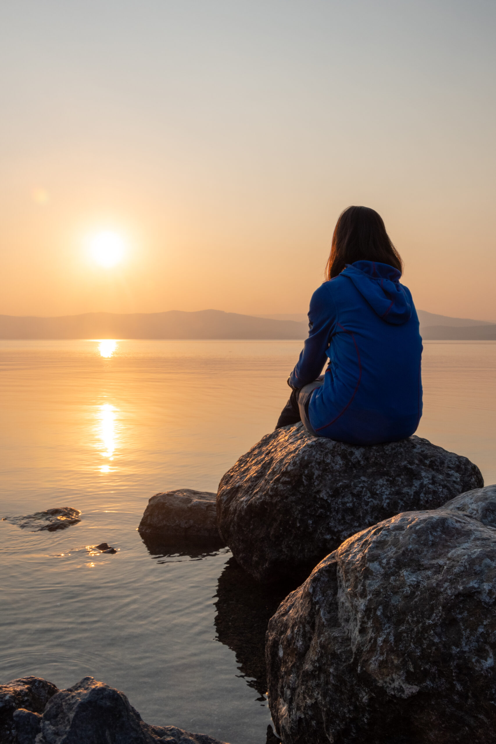 Person sitting on rock watching sunset over calm water.