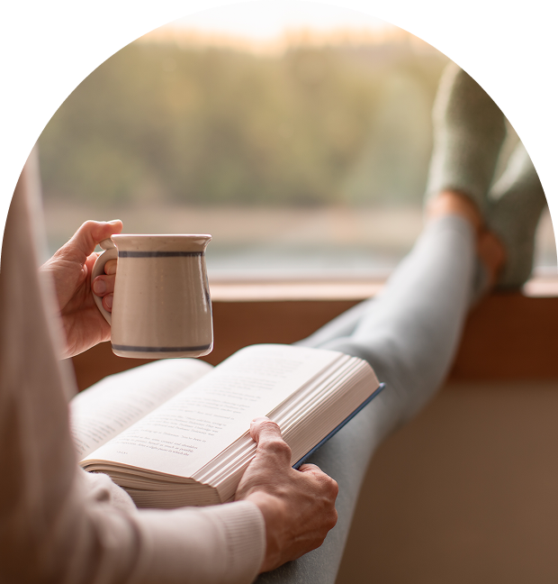 Person reading a book while holding a cup near a window.